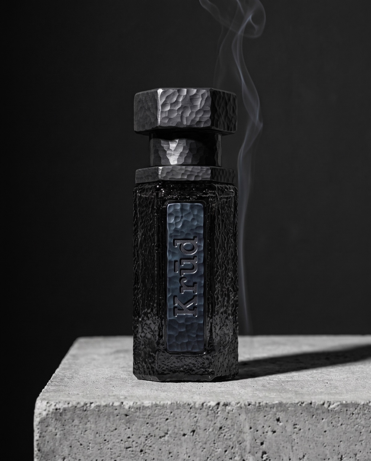 Noir — hand-hammered black glass bottle with iron hex cap, smoke rising against dark stone